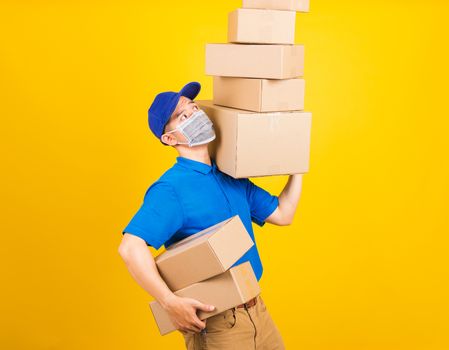 Asian young delivery worker man in blue t-shirt and cap uniform wearing face mask protective lifting stack a lot of boxes, under coronavirus or COVID-19, studio shot isolated yellow background