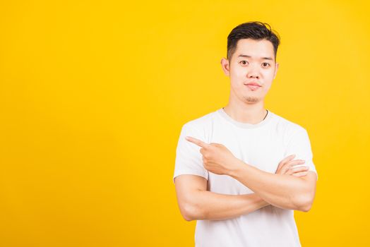 Portrait happy Asian handsome young man smiling standing wearing white t-shirt pointing finger to the side away he looking to camera, studio shot isolated yellow background