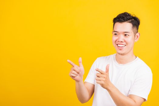 Portrait happy Asian handsome young man smiling standing wearing white t-shirt pointing finger to the side away he looking to space, studio shot isolated yellow background