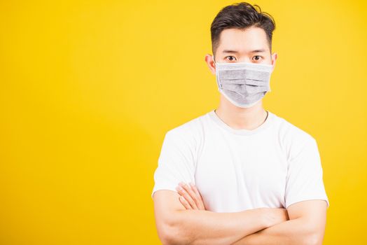 Closeup Asian young handsome man wearing face mask protective germ virus and stand crossed arm, studio shot isolated on yellow background and copy space, medical outbreak coronavirus COVID-19 concept