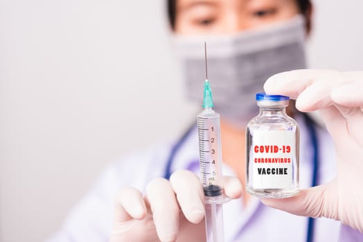 Asian female woman doctor or nurse in uniform and gloves wearing face mask protective in lab hold needle syringe drug and medicine vial vaccine bottle and on bottle has "COVID-19 VACCINE" text label