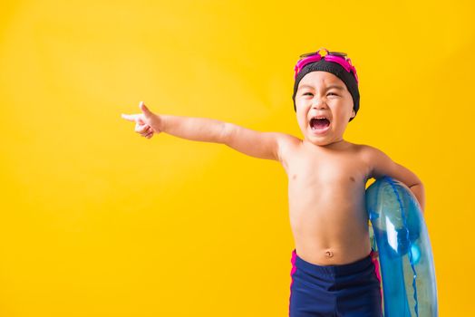 Summer vacation concept, Portrait Asian happy cute little child boy wear goggles and swimsuit hold blue inflatable ring, Kid hav fun point finger to side away, studio shot isolated yellow background