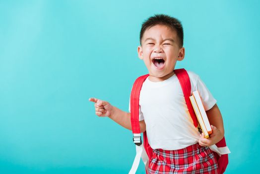 Back to school concept. Portrait Asian happy funny cute little child boy smile hug books and point finger to side away space, isolated blue background. Kid from preschool kindergarten with school bag