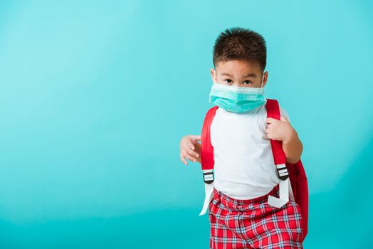 Back to school coronavirus Covid-19 education. Portrait Asian cute little child boy kindergarten wear face mask protective and school bag before going to school, studio shot isolated blue background