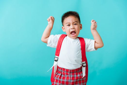 Back to school. Portrait happy Asian cute little child boy in uniform smile raise hands up glad when go back to school, isolated blue background. Kid from preschool kindergarten with school backpack