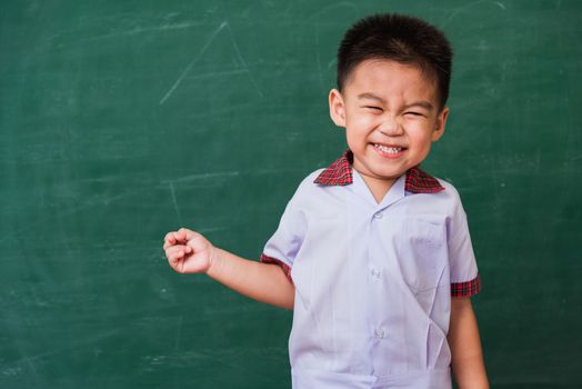 Back to School. Happy Asian funny cute little child boy from kindergarten in student uniform smiling point finger to side away space on green school blackboard, First time to school education concept