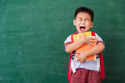 Back to School. Happy Asian funny cute little child boy from kindergarten in student uniform with school bag hold or hug books smile on green school blackboard, First time to school education concept