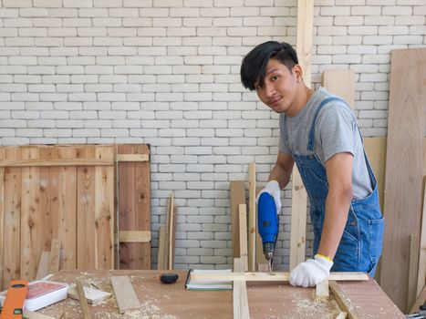 Asian carpentry uses an electric drill to drill the planks as they were prepared at the woodworking facility. Morning work atmosphere in the workshop room. A desk full of hand tools and wood piles.