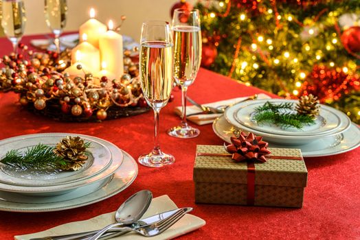 Beautifully decorated Christmas table - gift, glasses of champagne and tableware with Christmas decoration on a red tablecloth.