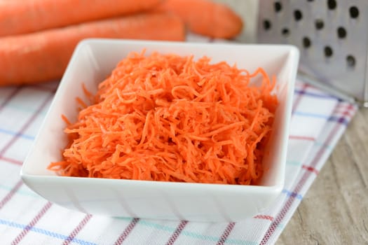 Concept of preparing a healthy salad - grated carrot in a white platter on a background of graters and carrots.