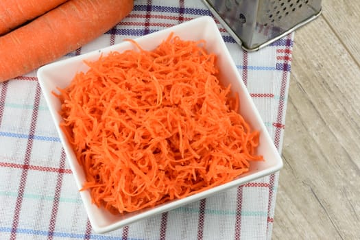 Concept of preparing a healthy salad - grated carrot in a white platter on a background of graters and carrots.