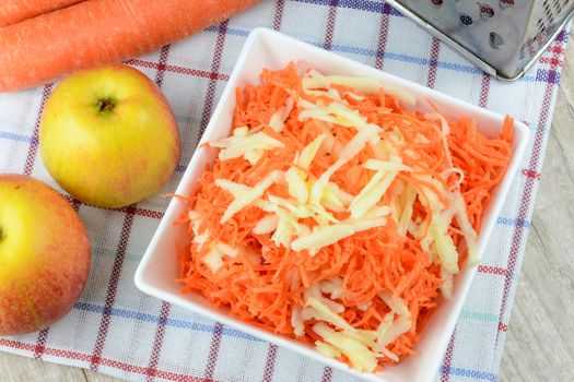 Concept of preparing a healthy salad - grated carrot with apple in a white platter on a background of graters, carrots and apples.