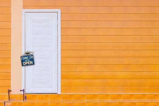 white wooden door and the orange wall with old rusty metalplate with word Come In We're Open haning on a knob, copyspace.