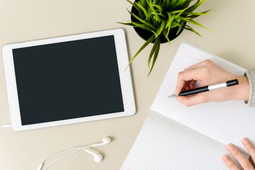 Concept image of student workplace - girl writing a homework in the notebook on the desk next to a digital tablet with blank screen, earphone and flower (high angel view).