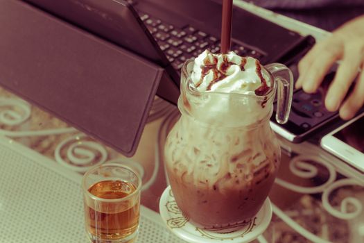 iced chocolate milk with whip cream on the background of  blurred hand using computer, business concept. Vintage retro style.