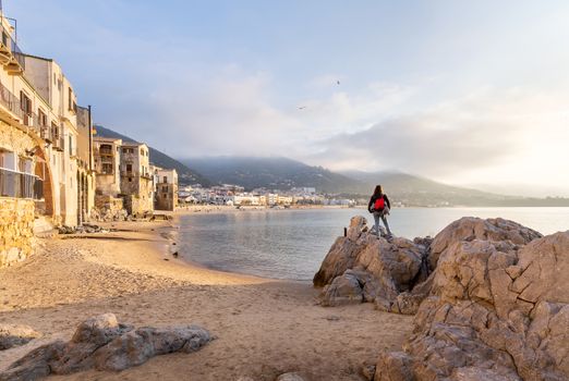Woman standing on a rock looking at the view of houses and long sandy beach in the old harbour on a sunny day in Cefalu, Sicily, Southern Italy.