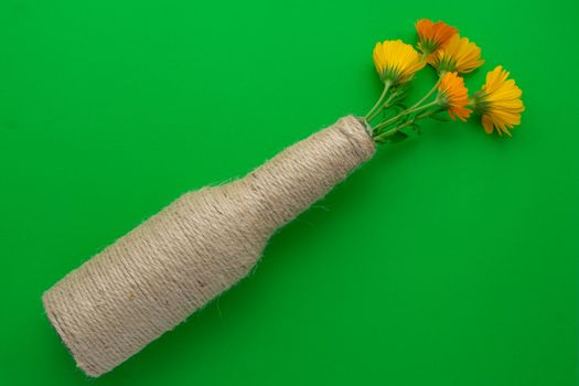 Glass beer bottle reused with hemp thread and flowers on the diagonal with a green background. Recycling and reusable concept