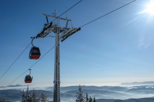 Gondola cabin lift in the ski resort over the forest on the background of snowy mountains in sunny day with copy space.