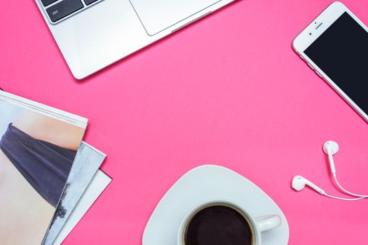 Concept of a creative workplace for a modern woman - a laptop, a smartphone with headphones, fashion catalogs and coffee on a pink desk (top view and copy space)