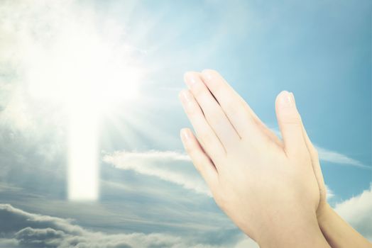 Concept of Christian prayer - folded hands for prayer against a blue sky and a shining cross sign (mixed)