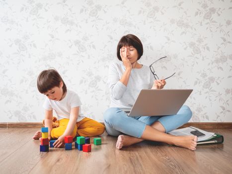 Mom and son sit at home quarantine because of coronavirus COVID19. Tired mother works remotely with laptop, son plays with toy blocks. Self isolation at home.