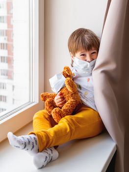 Toddler boy sits with teddy bear in medical masks. Kid with plush toy plays hide-and-seek behind curtains. Browh plush bear and child on home quarantine because of coronavirus COVID-19.