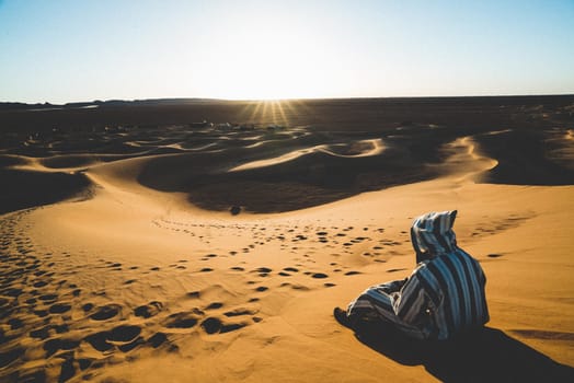 Arab person with black and white jellaba sit in the sand sees the sahara desert and a near camp