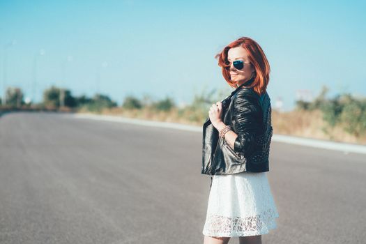 red-haired girl in a black jacket and blue glasses on a gray road