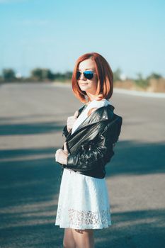 red-haired girl in a black jacket and blue glasses on a gray road