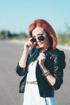 red-haired girl in a black jacket and blue glasses on a gray road