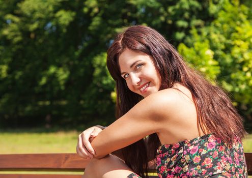 portrait of young beautiful smiling brunette sitting on a bench in a city park on sunny summer day closeup