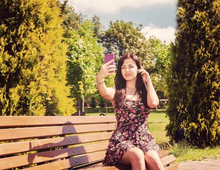 young girl making selfie sitting on a park bench on sunny summer day
