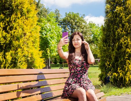young beautiful girl making selfie sitting on a park bench on sunny summer day