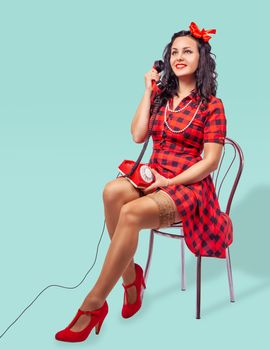 smiling young pinup woman in red dress and nylon stockings sitting on a chair and talking on phone over green background