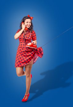 happy young pinup standing woman in red dress and nylon stockings talking 
on phone over blue background