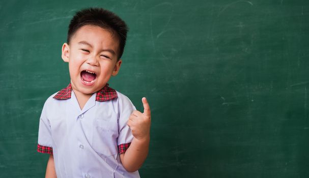 Back to School. Happy Asian funny cute little child boy from kindergarten in student uniform smiling point finger to side away space on green school blackboard, First time to school education concept