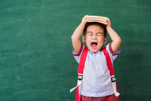 Back to School. Happy Asian funny cute little child boy from kindergarten in student uniform with school bag and book on head smiling on green school blackboard, First time to school education concept