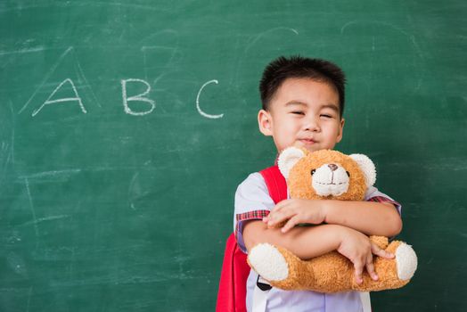 Back to School. Happy Asian funny cute little child boy from kindergarten in student uniform with school bag smiling and hugging teddy bear on green school blackboard, First time to school education