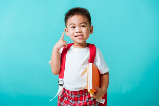 Back to school concept. Portrait Asian happy funny cute little child boy smile hug books and point finger to side away space, isolated blue background. Kid from preschool kindergarten with school bag