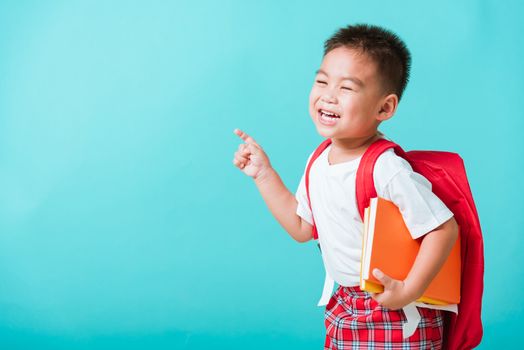 Back to school concept. Portrait Asian happy funny cute little child boy smile hug books and point finger to side away space, isolated blue background. Kid from preschool kindergarten with school bag