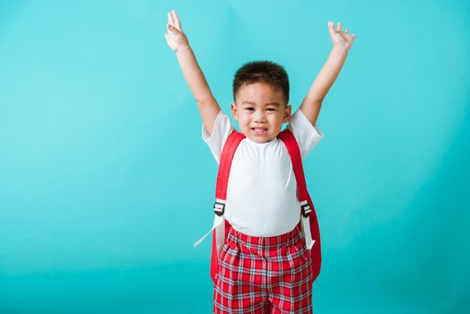 Back to school. Portrait happy Asian cute little child boy in uniform smile raise hands up glad when go back to school, isolated blue background. Kid from preschool kindergarten with school backpack