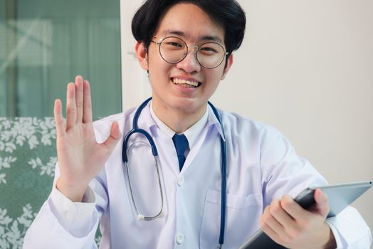 Happy Asian young doctor handsome man smile in uniform wear glasses raise hands to greet patients modern smart digital tablet computer on desk at hospital office, Technology Healthcare And Medicine