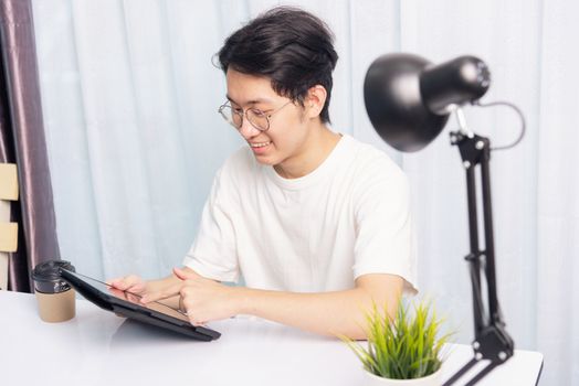 Happy Asian young business handsome man work from home office wear glasses, t-shirt comfortable he smiling and using a black modern smart digital tablet computer to read email or learning on desk