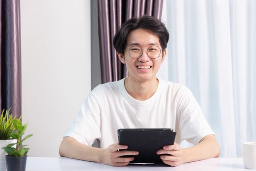 Happy Asian young business handsome man work from home office wear glasses, t-shirt comfortable he smiling and using a black modern smart digital tablet computer to read email or learning on desk