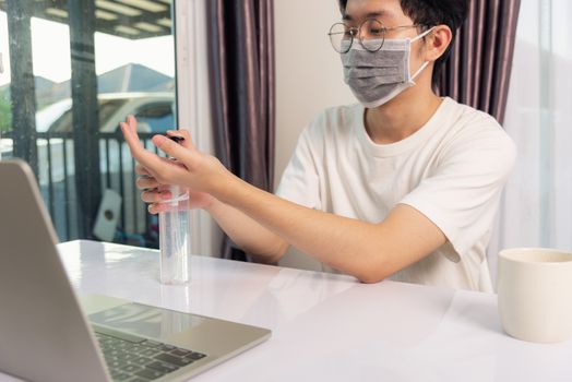 Asian business young handsome man glasses working from home office he quarantines disease coronavirus or COVID-19 wearing protective mask and cleaning hands with sanitizer gel on front laptop computer