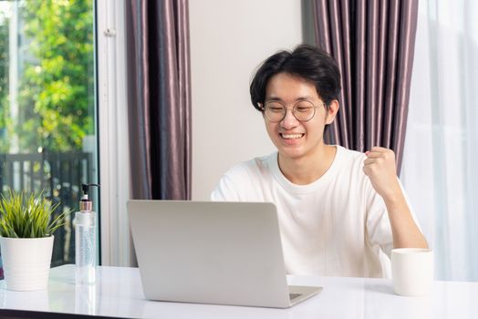 Happy Asian young business handsome man smile work from home office wear glasses, t-shirt comfortable exciting good news successful winner raises hand yes gesture with laptop computer on desk at home