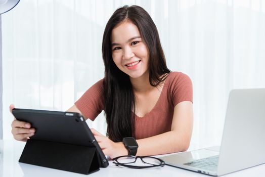 Work from home, Smiling Happy Asian business young beautiful woman sitting on desk workspace use modern smart tablet and laptop computer technology video call conferencing to team at home office