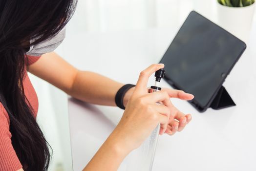 Asian Business young woman working from home office he quarantines disease coronavirus or COVID-19 wearing a protective mask and cleaning hands with sanitizer gel on front tablet computer