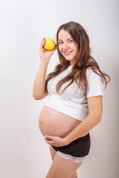 Image of pregnant woman touching her big belly and holding yellow apple in the hand on white background. Close up. Beautiful body of pregnant woman. Motherhood, pregnancy. Woman expecting baby