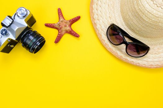 Summer holidays and vacation concept - sunglasses, starfish and a beach straw hat next to retro slr camera on a yellow background with copy space.